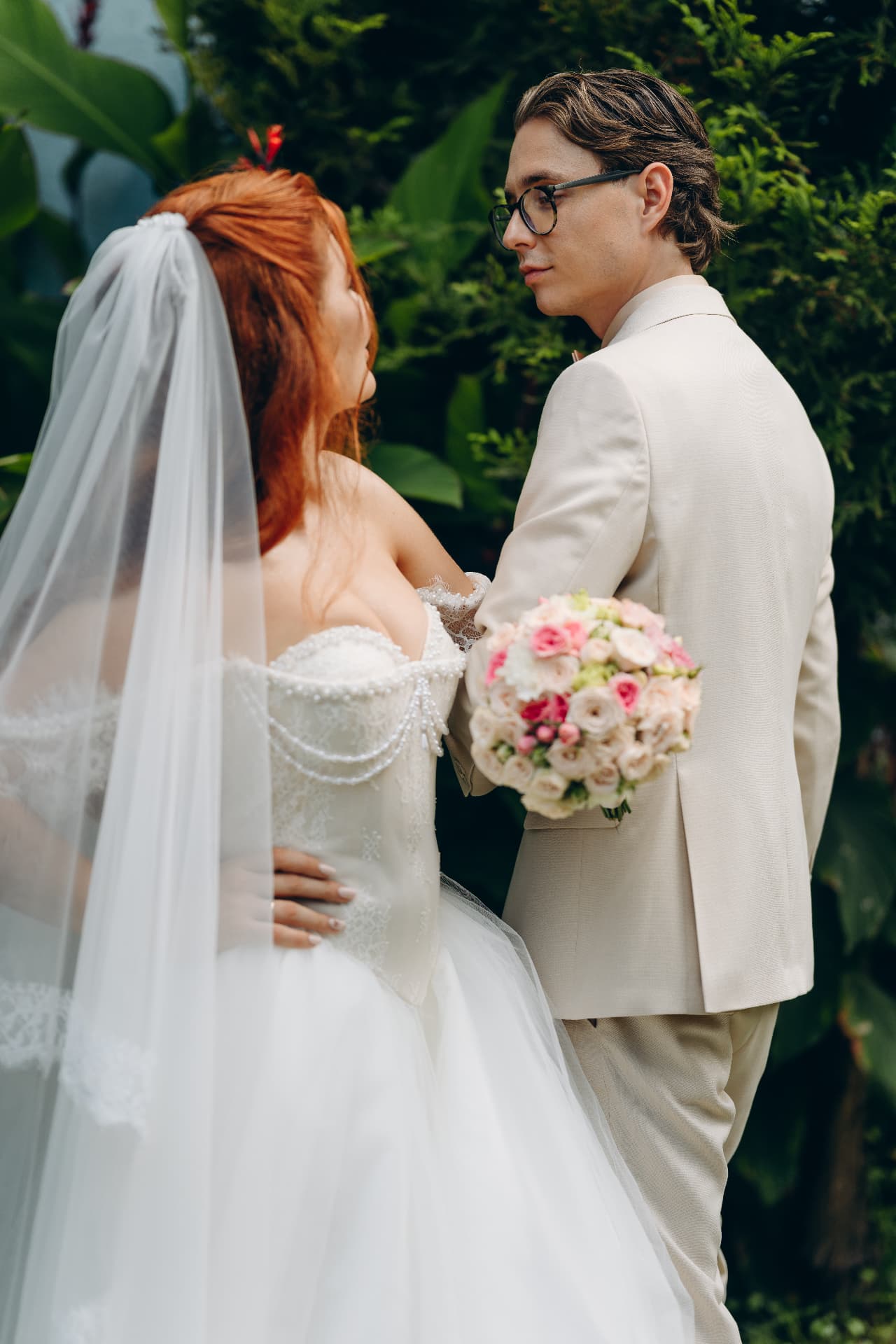 Bride and groom in cream suit among tropical greenery with a pink and cream bouquet