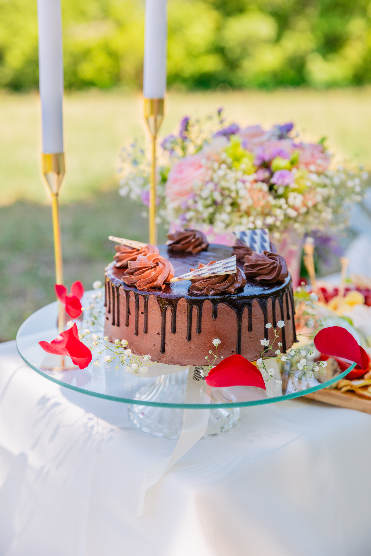 Premium chocolate drip cake on crystal stand with rose petals — luxury Budapest picnic by Vivielle Events