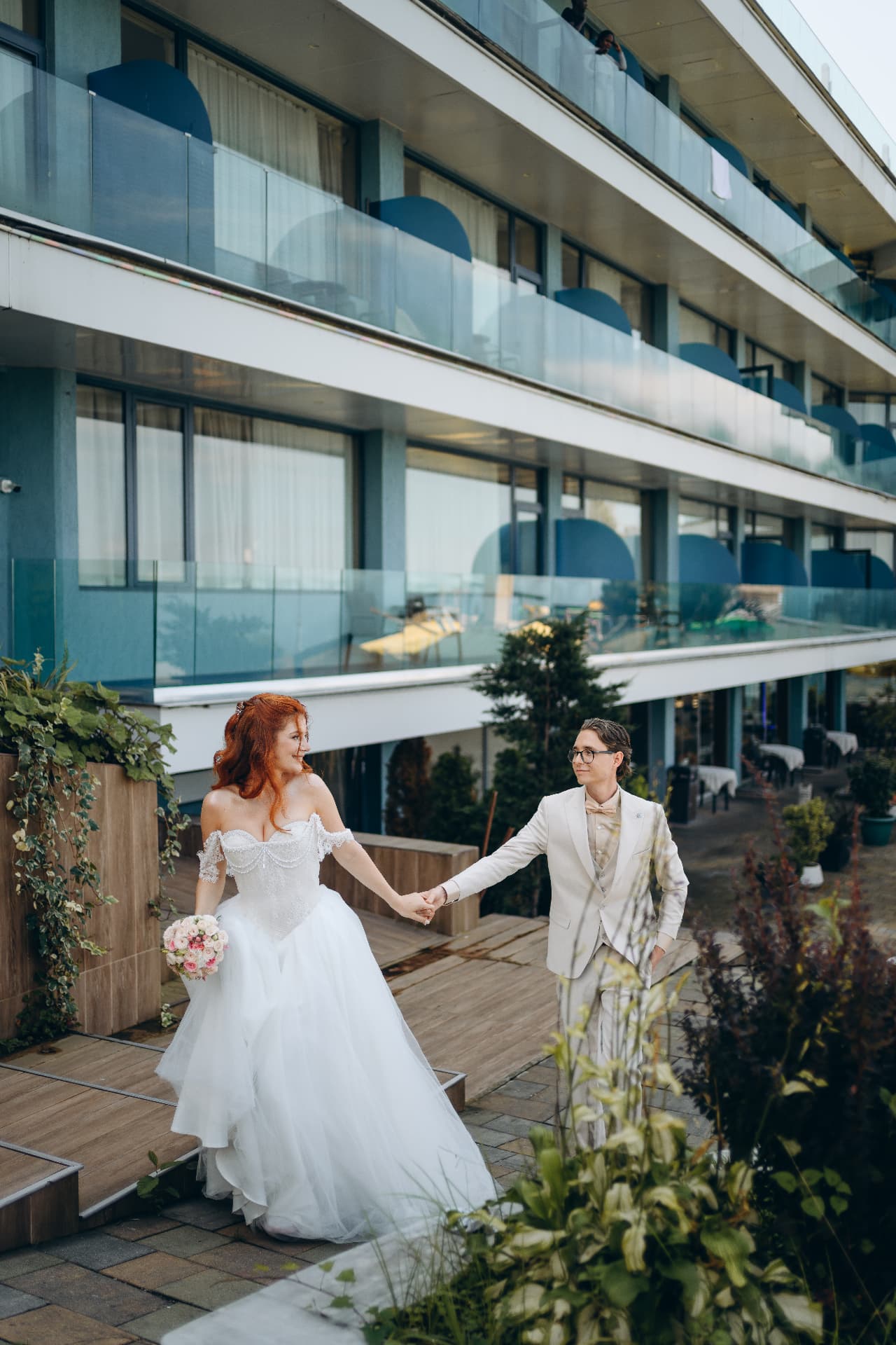 Bride and groom holding hands walking along a terrace at a modern waterfront venue