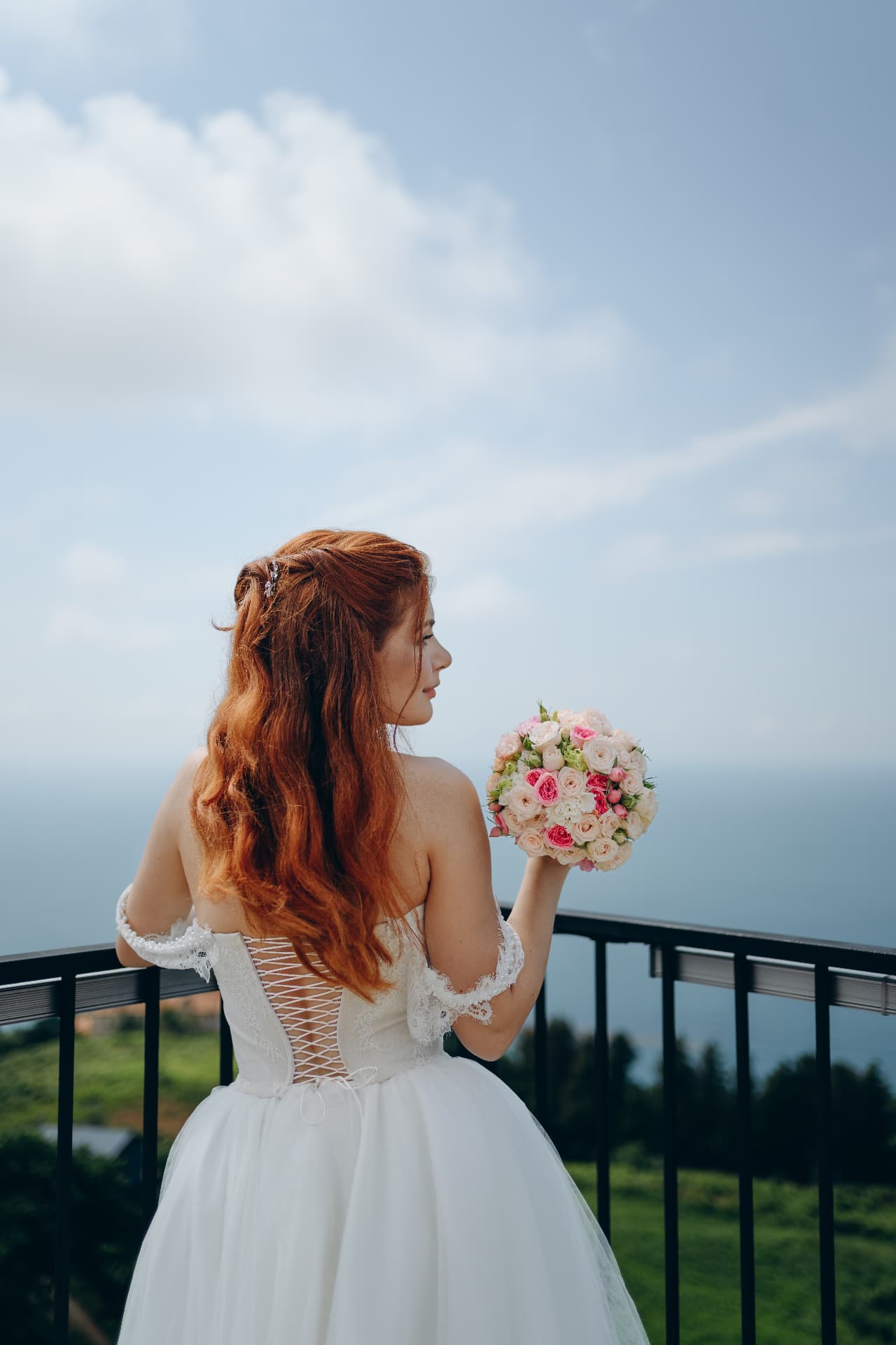 Bride from behind holding her bouquet, looking out over the sea from a balcony